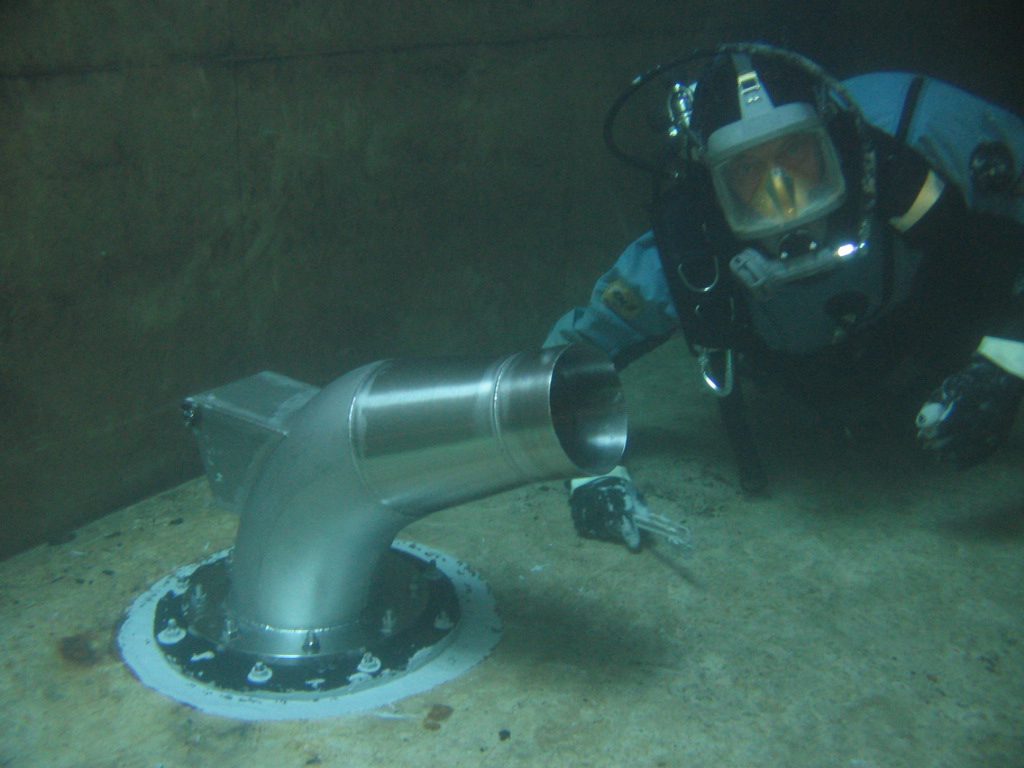 A diver looking at the camera after sealing around the base of a stainless steel nozzle in a concrete tank.
