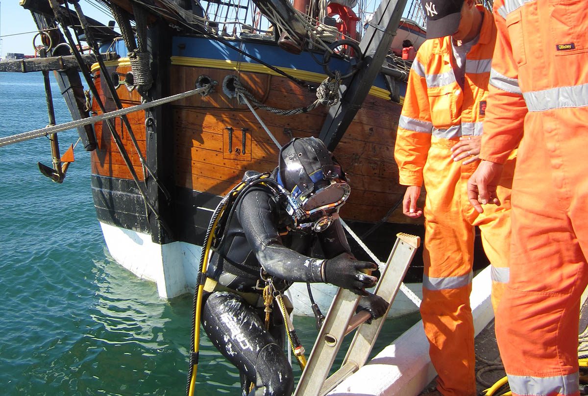 A diver climbing down the pier ladder to enter the water before conducting repairs on the wooden hull of the ship.