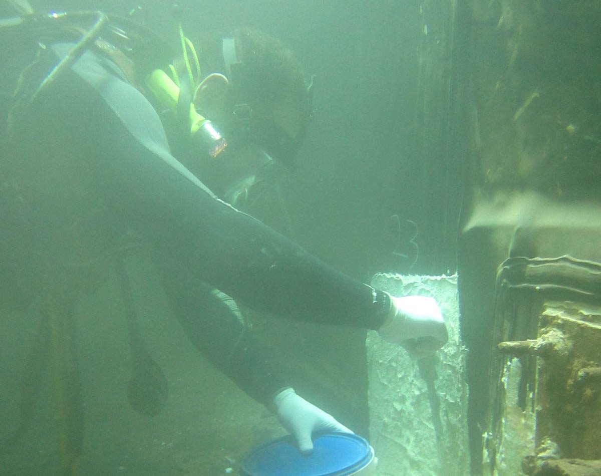 A diver using a spatula to apply Scubapoxy 1725 onto the floor and sides of the tank.