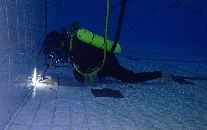 A diver underwater in the swimming pool using a caulking gun to apply the new sealant into the expansion joints.