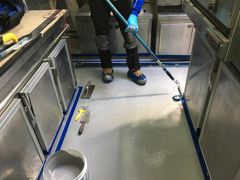 An installer using a roller to apply an industrial epoxy floor coating in a commercial kitchen.