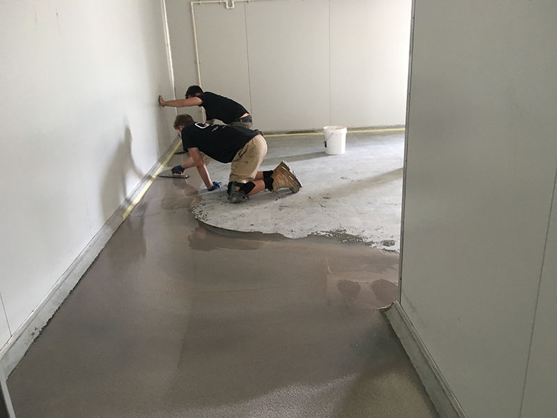 Two installers using trowels to re-level the floor with a screed before applying an industrial epoxy floor in a factory.