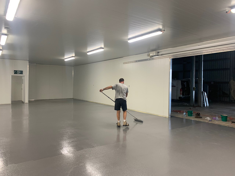 An installer using a roller to apply two coats of industrial epoxy floor coating in a warehouse.