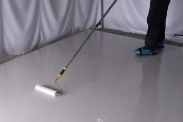 An installer uses a roller tray to apply the topcoat on a light grey, protective epoxy flooring system.